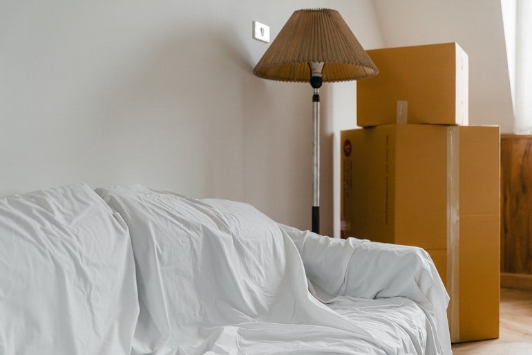 A couch covered with white sheets, with a lamp and some big boxes on the side. Big couches can be one of the most difficult furniture to move.