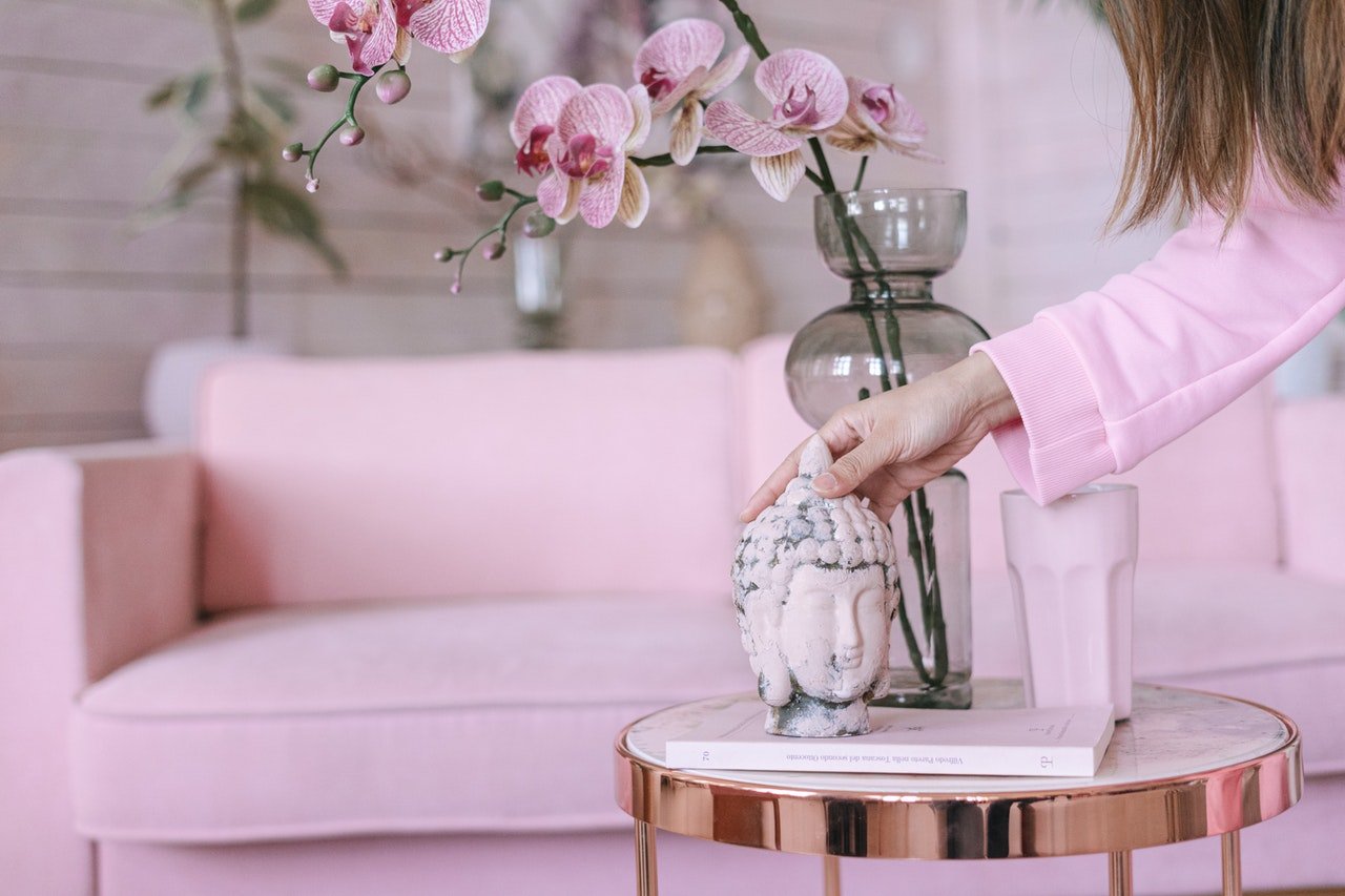 A woman in pink sets up a sculpture next to an orchid with a pink sofa in the background.