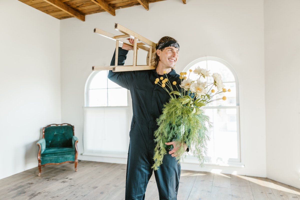 Man is jumpsuit carrying a wooden stool and flower n