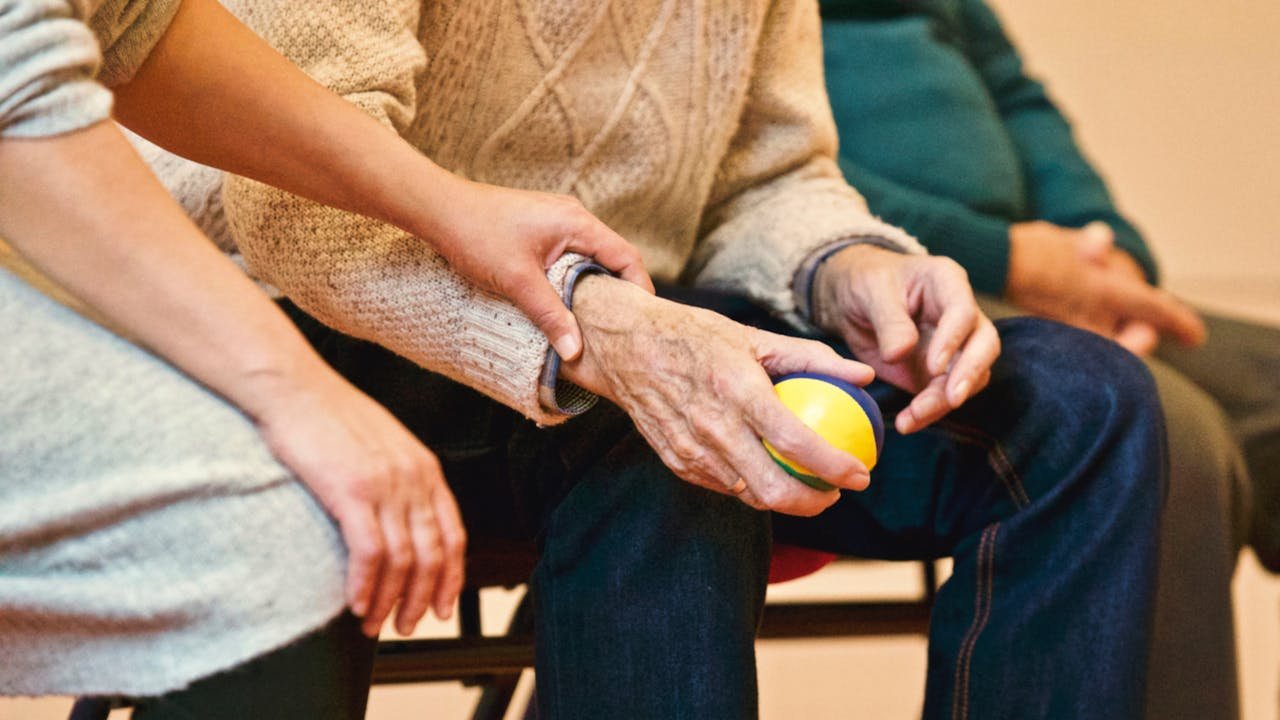 An elderly person's hand holding a colorful ball, with a caregiver's hand gently resting on their wrist, conveying support and care.
